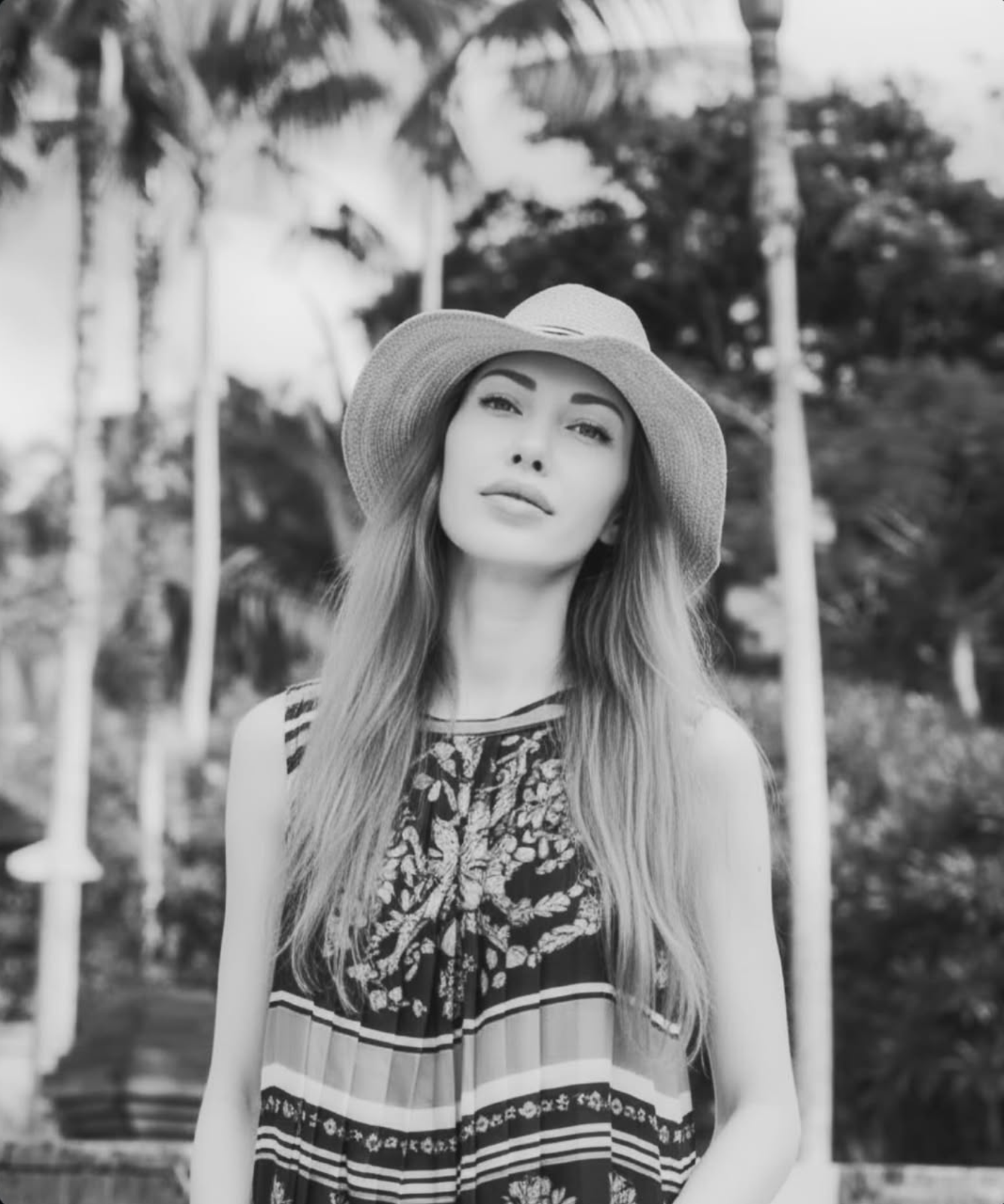 Sarah Marks wearing a wide-brimmed hat and patterned sleeveless dress outdoors in a tropical setting.