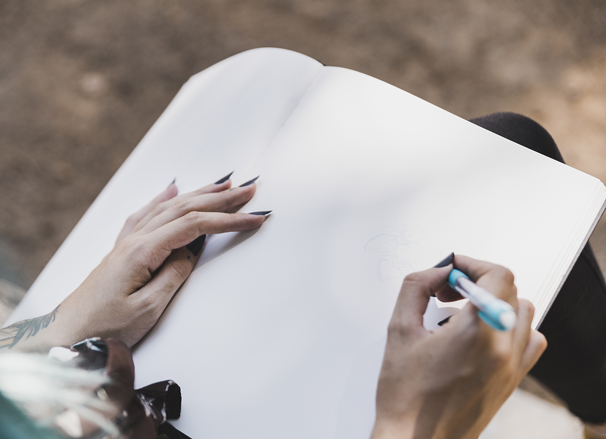 Person with black nails sketching a flower on a blank notebook page outdoors.