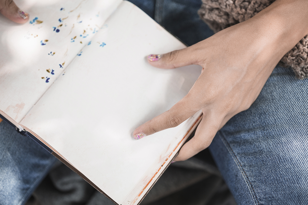 A person with decorated nails holding open a blank page of a notebook while wearing jeans.