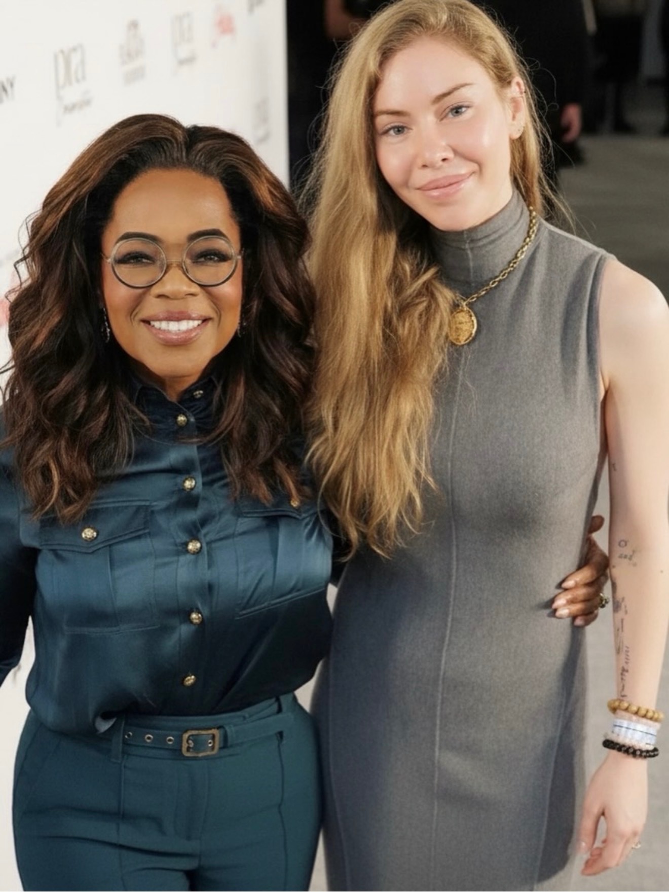 Two women smiling Sarah Marks and Oprah Winfrey at an event with a red carpet backdrop and lights overhead.