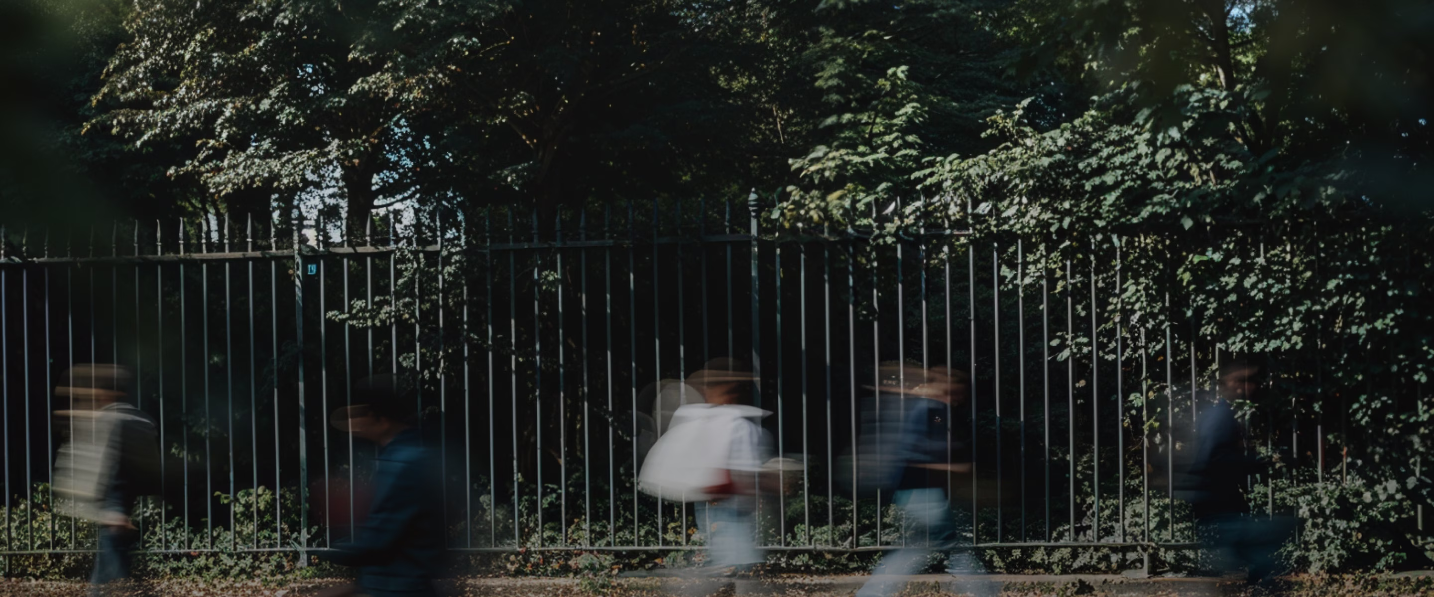 Personnes floues marchant devant une clôture métallique avec des arbres et des feuillages en arrière-plan.