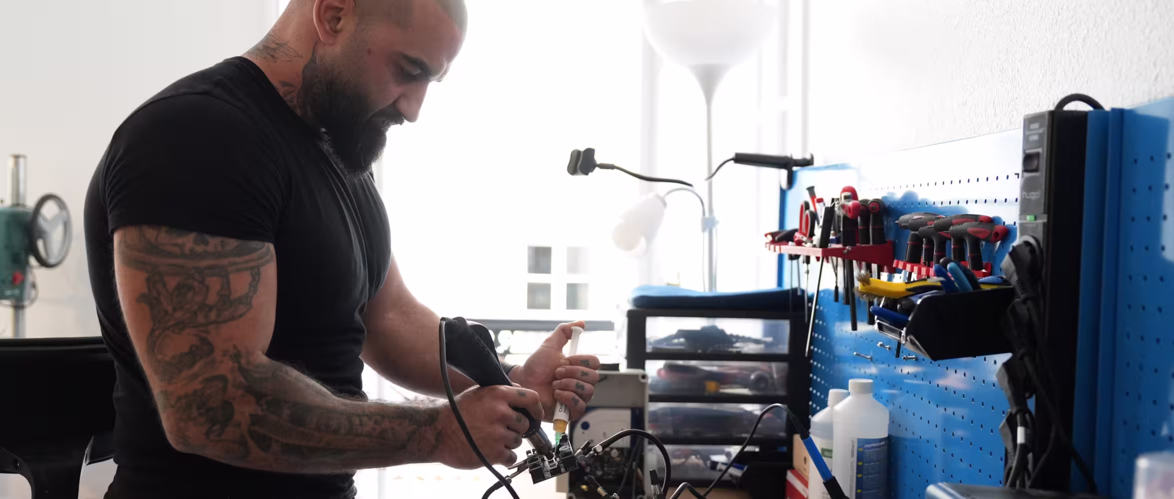 Homme tatoué utilisant un fer à souder pour réparer un circuit électronique dans un atelier avec outils accrochés au mur bleu.