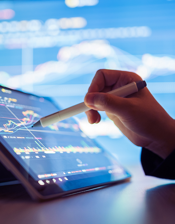 Hand using a stylus on a digital tablet displaying financial graphs and charts, with a blurred screen of data in the background.