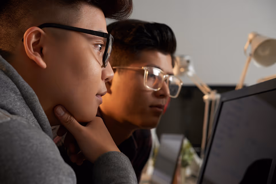 Two young men wearing glasses closely looking at a computer screen in a dimly lit room.