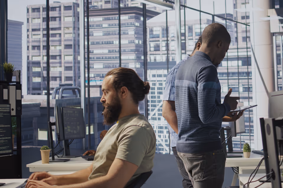 Bearded man with hair tied back working at a desktop computer in a modern office with large windows, while two colleagues stand nearby discussing and looking at a tablet.