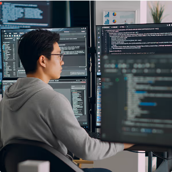 Man wearing glasses and a gray hoodie working on multiple monitors displaying code and data in a tech workspace.