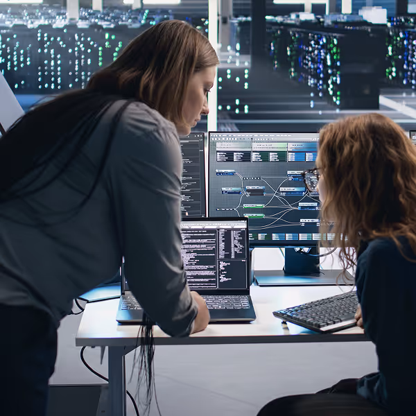 Two women working together at a desk with a laptop and dual monitors showing complex data and code in a futuristic office.