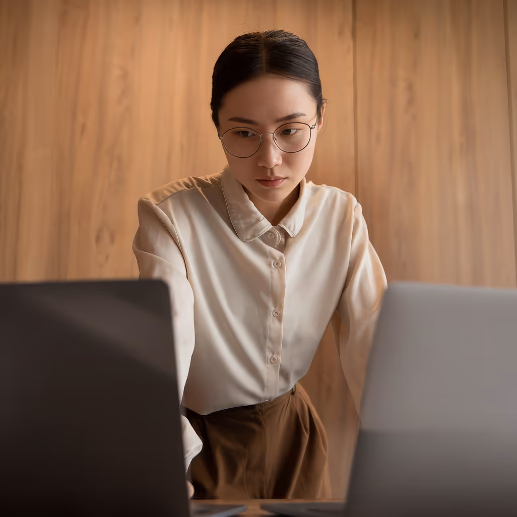 Woman wearing glasses focused on work between two laptops with wooden wall background.