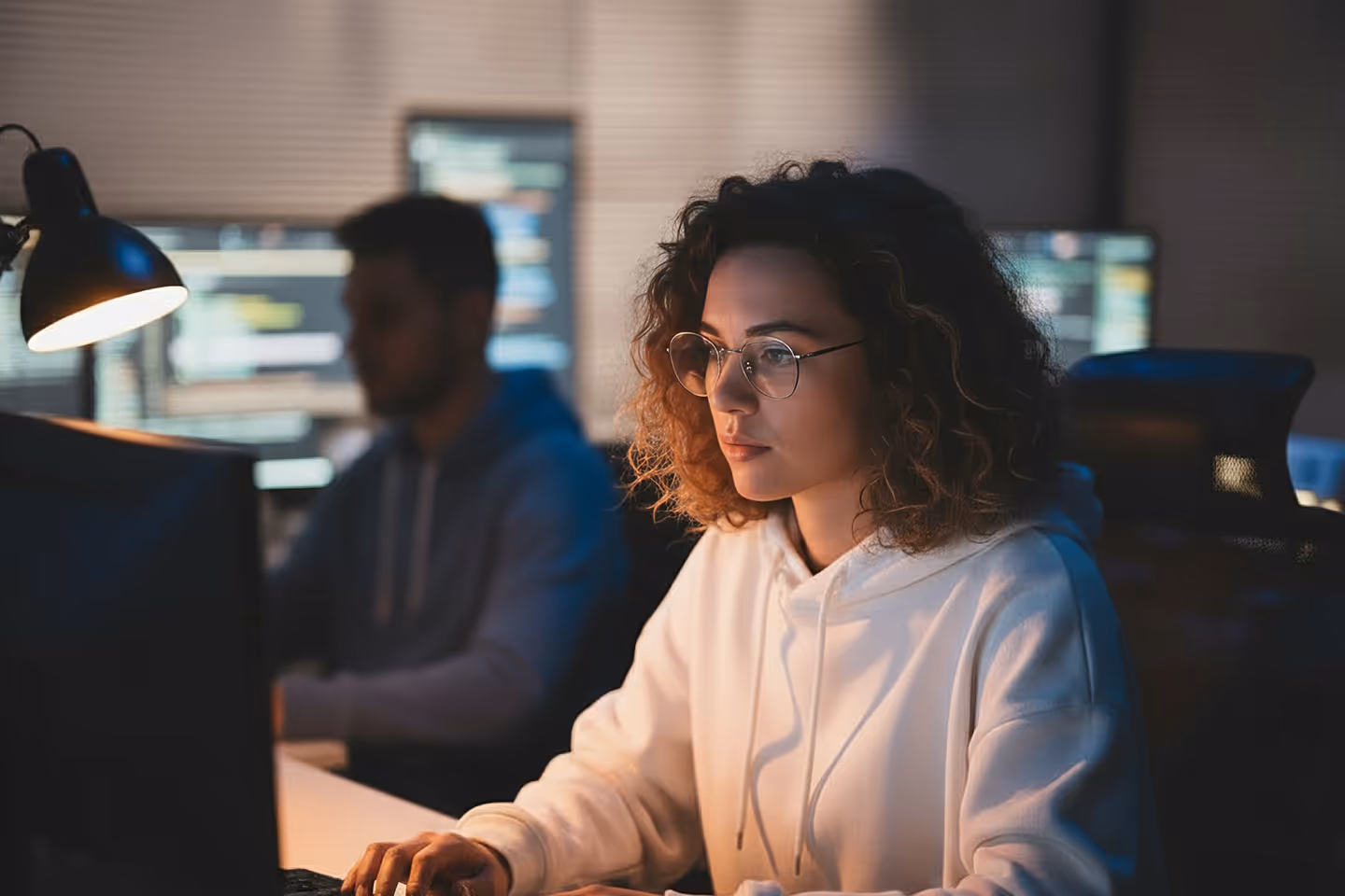 Focused woman with curly hair and glasses working on a computer in a dimly lit office with code on monitors behind her.