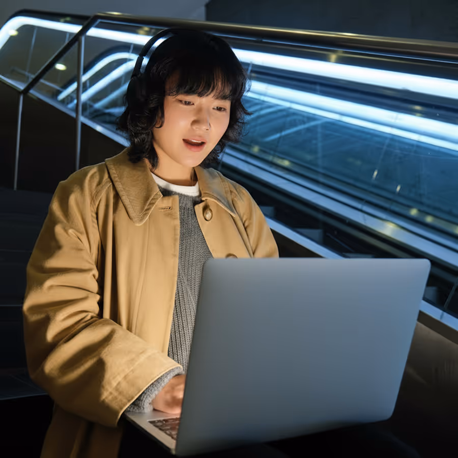 Young woman wearing headphones and beige coat using a laptop on stairs with escalator in the background.