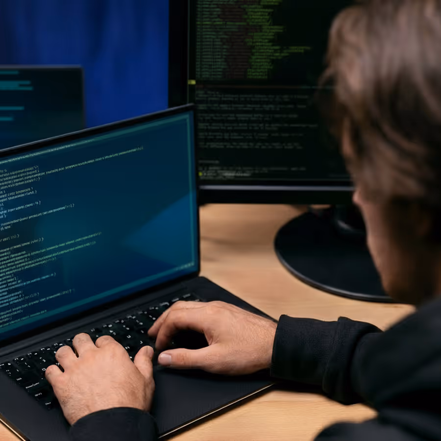 Person typing code on a laptop with multiple monitors displaying programming code.