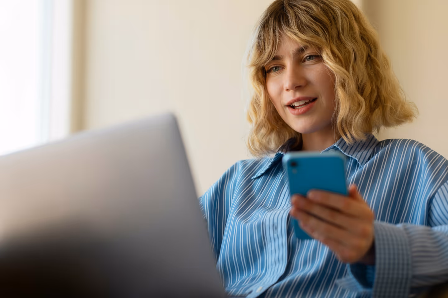Young woman in a blue striped shirt using a smartphone and laptop simultaneously indoors.