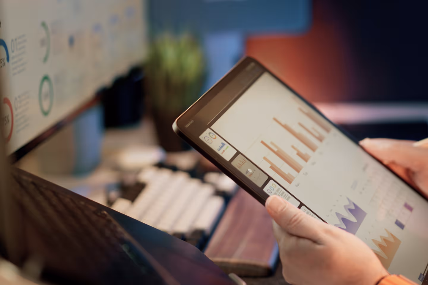 Person holding a tablet displaying various colorful charts and graphs, with a blurred computer monitor and keyboard in the background.