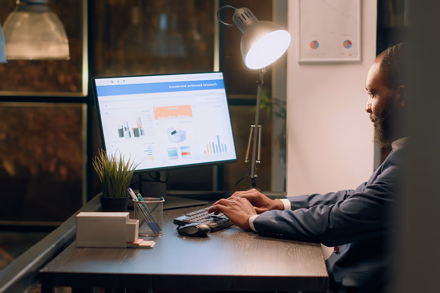 Man in a suit working at a desk with a computer displaying financial charts and graphs under a desk lamp.