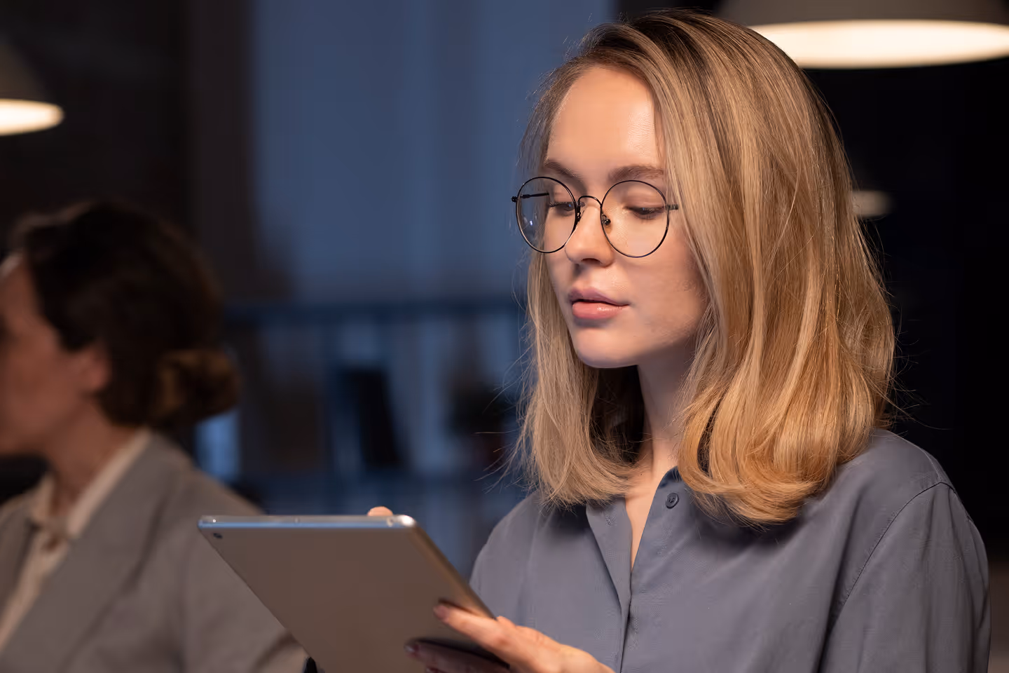 Young woman with glasses focused on a tablet in a dimly lit room.