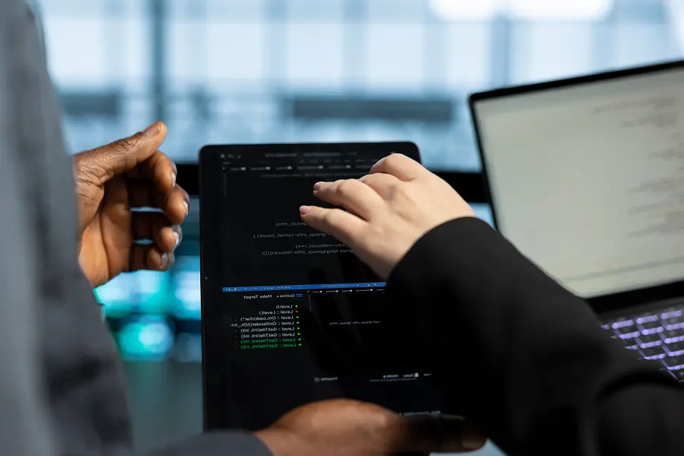 Two people interacting with a tablet displaying programming code, with a laptop in the background.