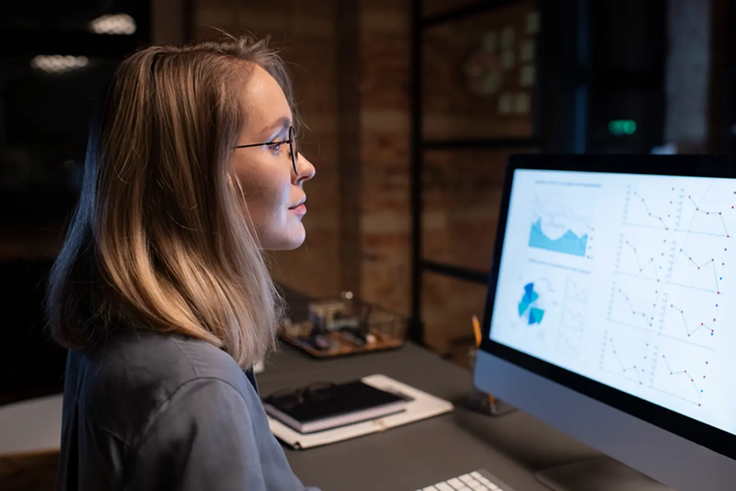 Woman with blonde hair and glasses analyzing charts and graphs on a large desktop computer screen in a dimly lit office.