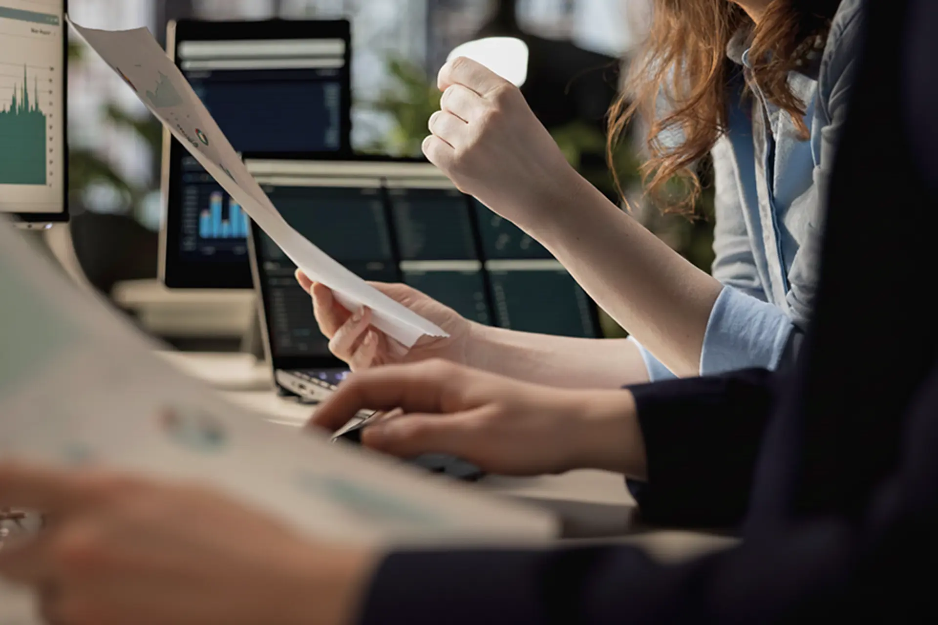 Close-up of two people analyzing printed charts and data on computer screens in an office setting.