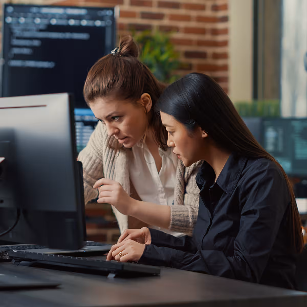 Two women collaborating and looking intently at a computer screen in an office with multiple monitors.