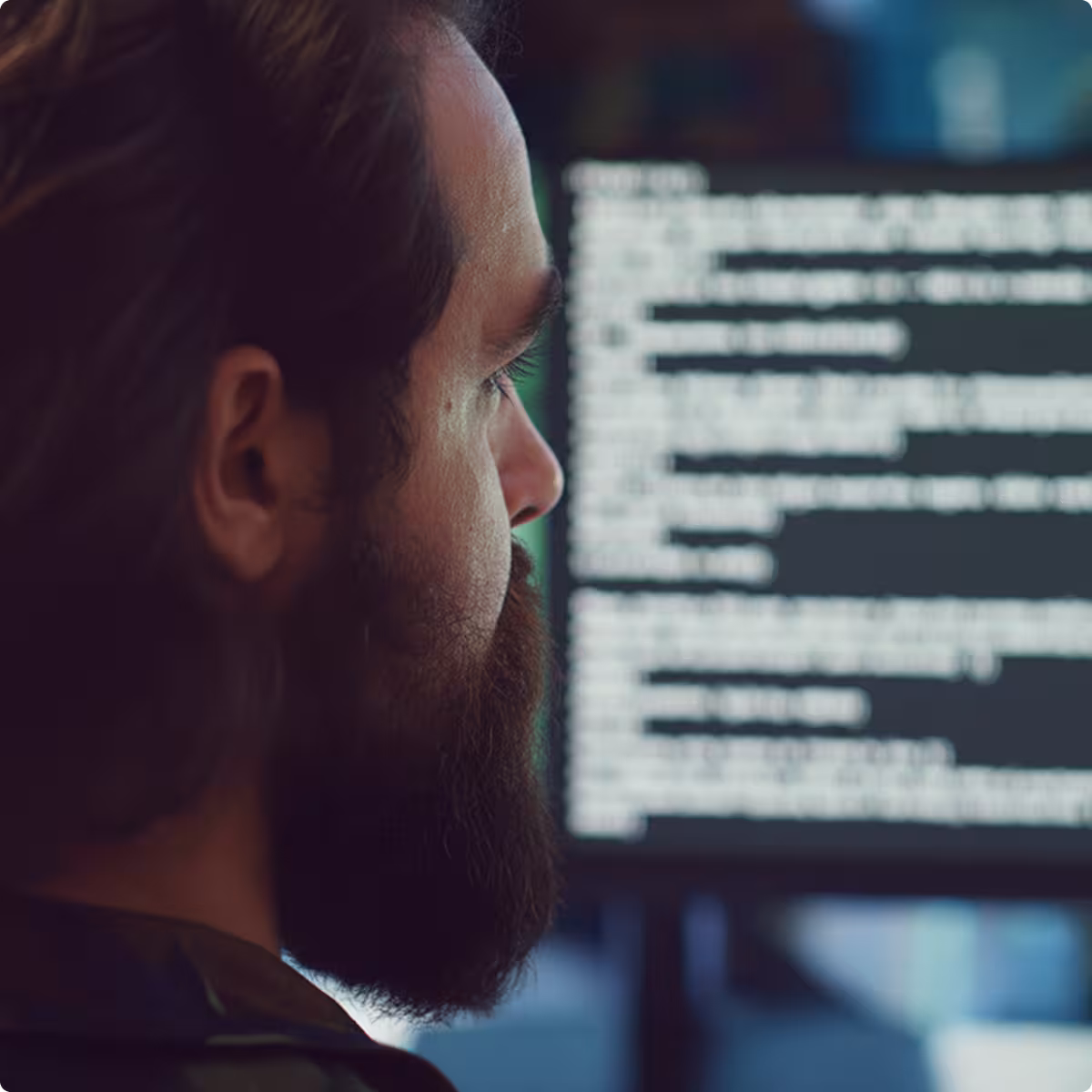 Man with a beard and long hair closely looking at a computer screen displaying blurred code.