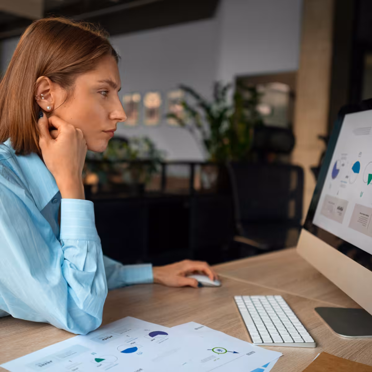 Woman in blue shirt intently analyzing charts on a computer screen and printed documents in an office.
