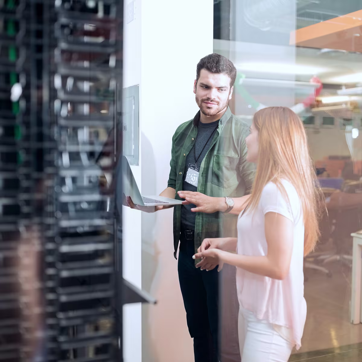 Two IT professionals, a man holding a laptop and a woman, discussing near a server rack in an office.