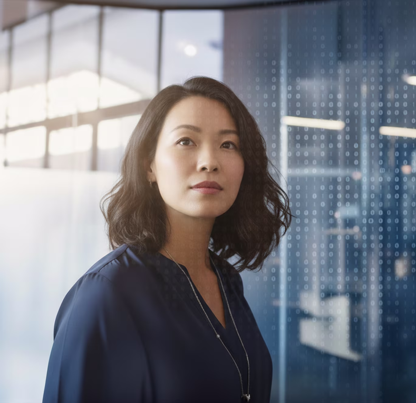 Confident woman with shoulder-length dark hair wearing a navy blouse and necklace, standing indoors with digital binary code overlay.