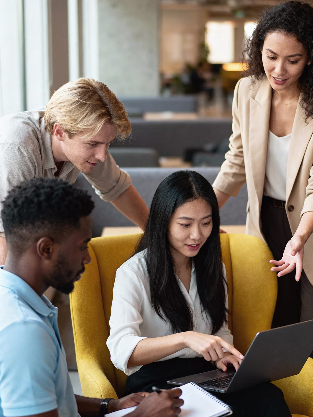 Four diverse colleagues collaborating around a laptop in a modern office lounge.