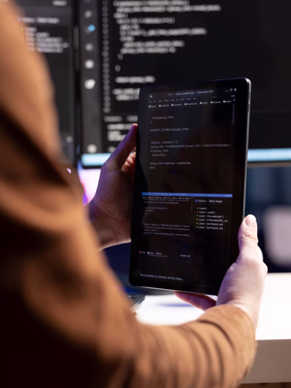 Person holding a tablet displaying code with a computer screen showing code in the background.