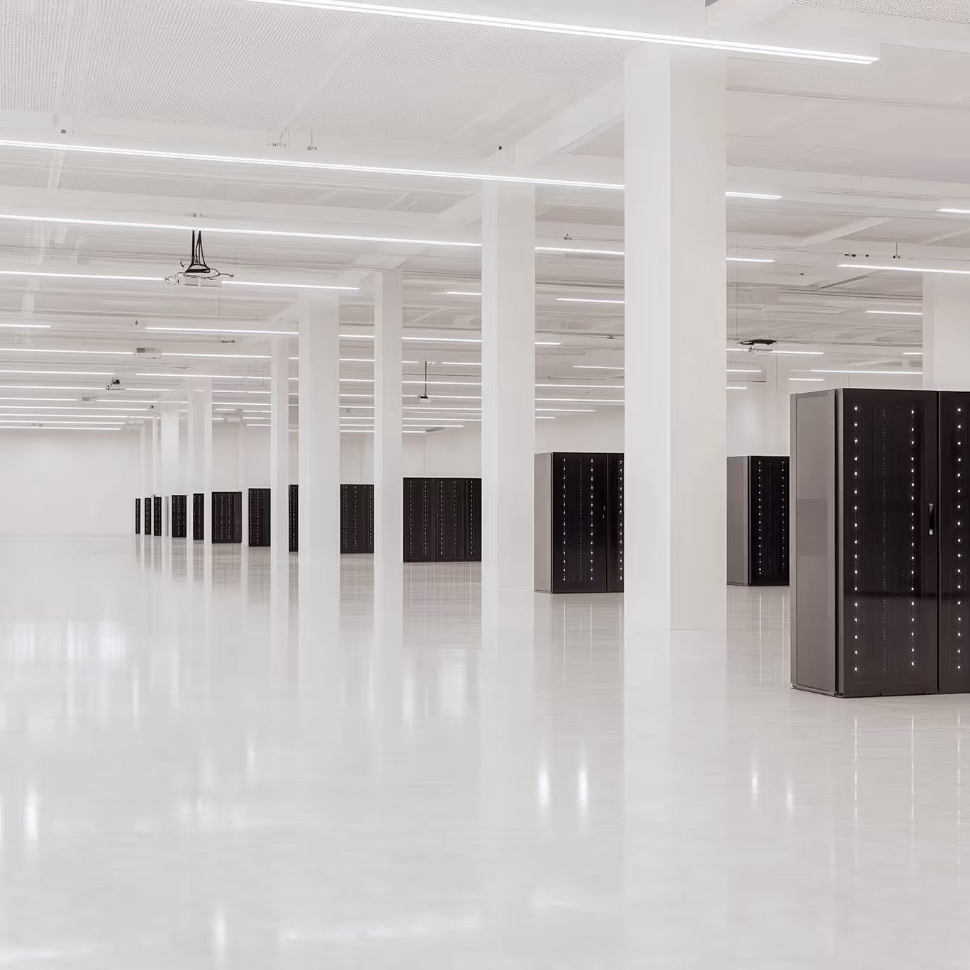 Large, empty data center room with rows of black server racks and white pillars under bright LED ceiling lights.