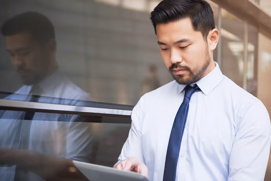 Man in white shirt and blue tie using a tablet while standing next to a glass wall reflecting his image.