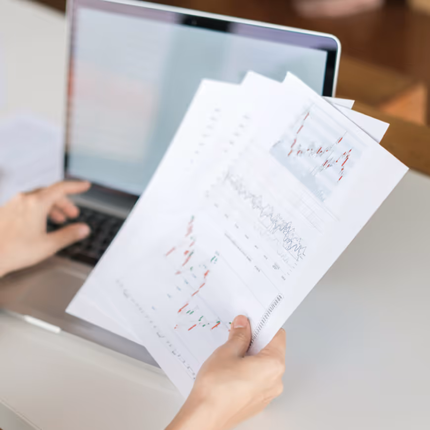 Person holding printed financial charts in front of a laptop keyboard.