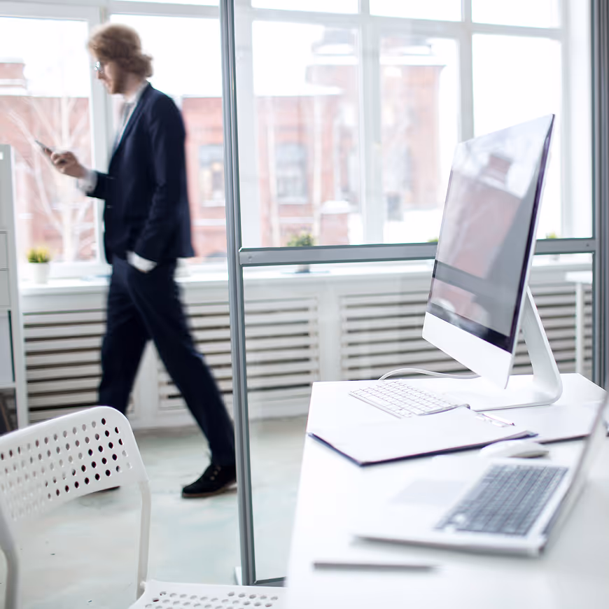 Modern office workspace with a desktop computer, laptop, and man in a suit walking while looking at his phone.
