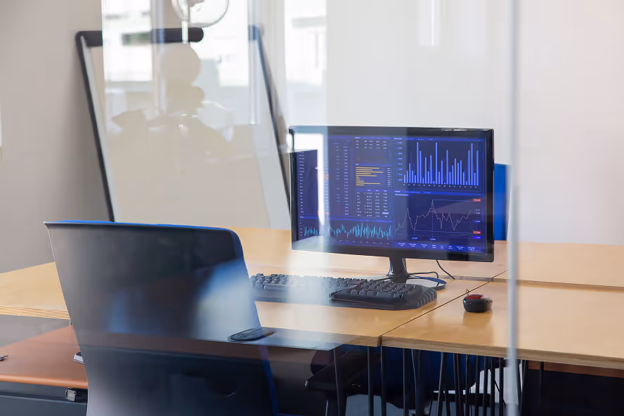 Office desk with a computer monitor displaying data charts and graphs, a keyboard, and a mouse, with a black office chair visible.