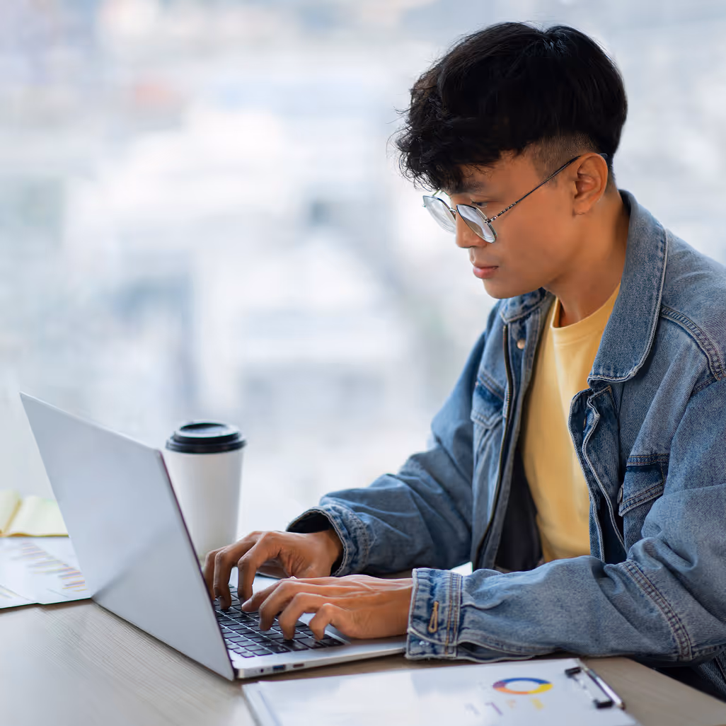 Young man wearing glasses and a denim jacket working on a laptop at a desk with a coffee cup and documents.