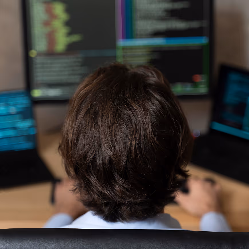 Person with dark hair working on a computer with multiple screens displaying code and technical data.