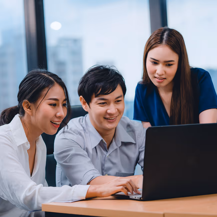 Three young professionals smiling and collaborating while looking at a laptop in an office with large windows.