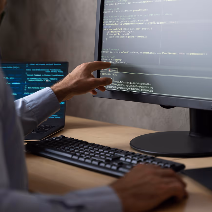 Person pointing at code on a computer monitor with a laptop and keyboard on the desk.