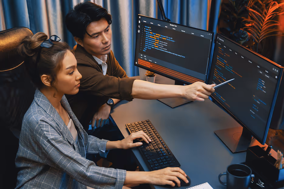 Two people collaborating at a desk with dual monitors displaying code, one pointing at the screen and the other typing on a keyboard.