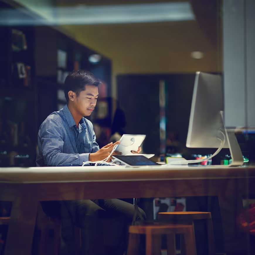 Man in a striped shirt working at a desk with a tablet and a computer in a dimly lit office.