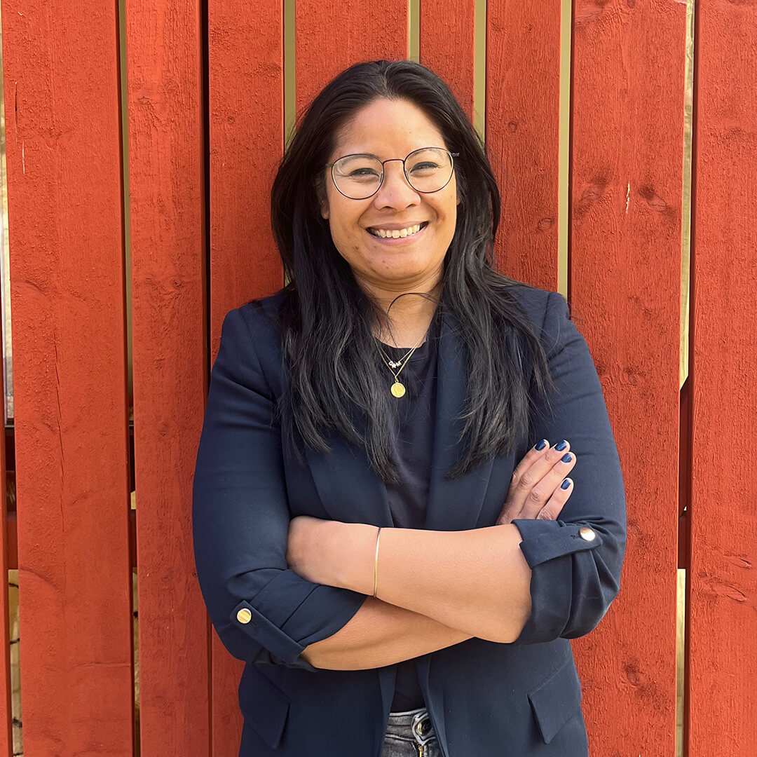 Person standing in front of a red wooden wall, wearing glasses and a dark blazer, smiling with arms crossed.