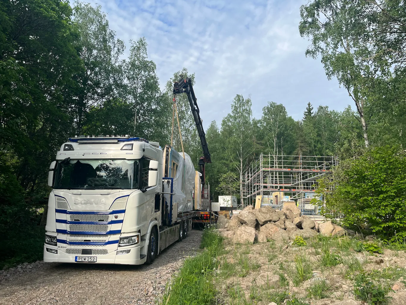 A white truck delivering building materials at a forest construction site, where scaffolding and building frames are visible among trees and rocks.
