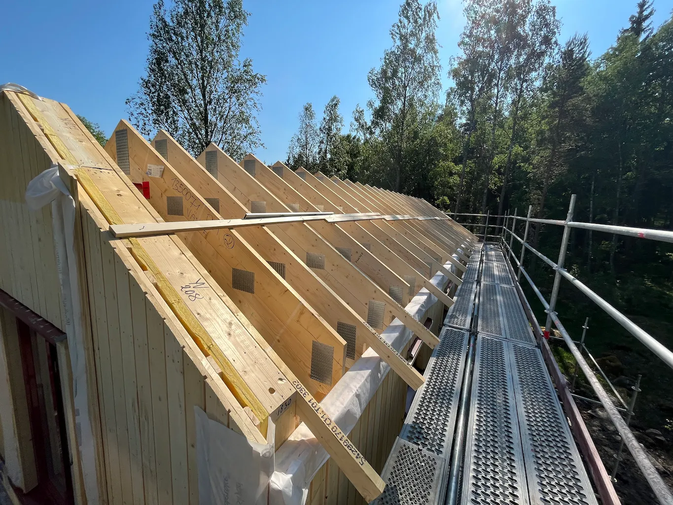 A wooden house under construction with exposed roof trusses, seen from a scaffolding platform under a clear blue sky, surrounded by tall trees