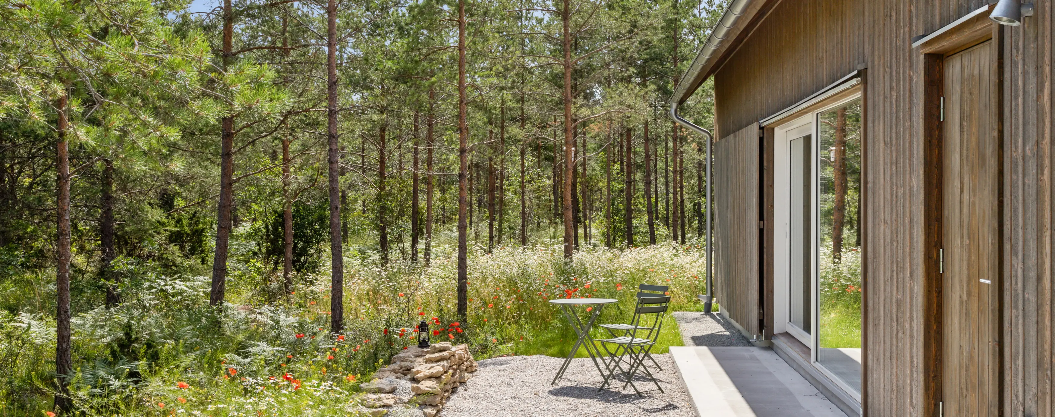 Minimalist outdoor seating area with a green metal table and chairs beside a wooden house, surrounded by wildflowers and pine trees.