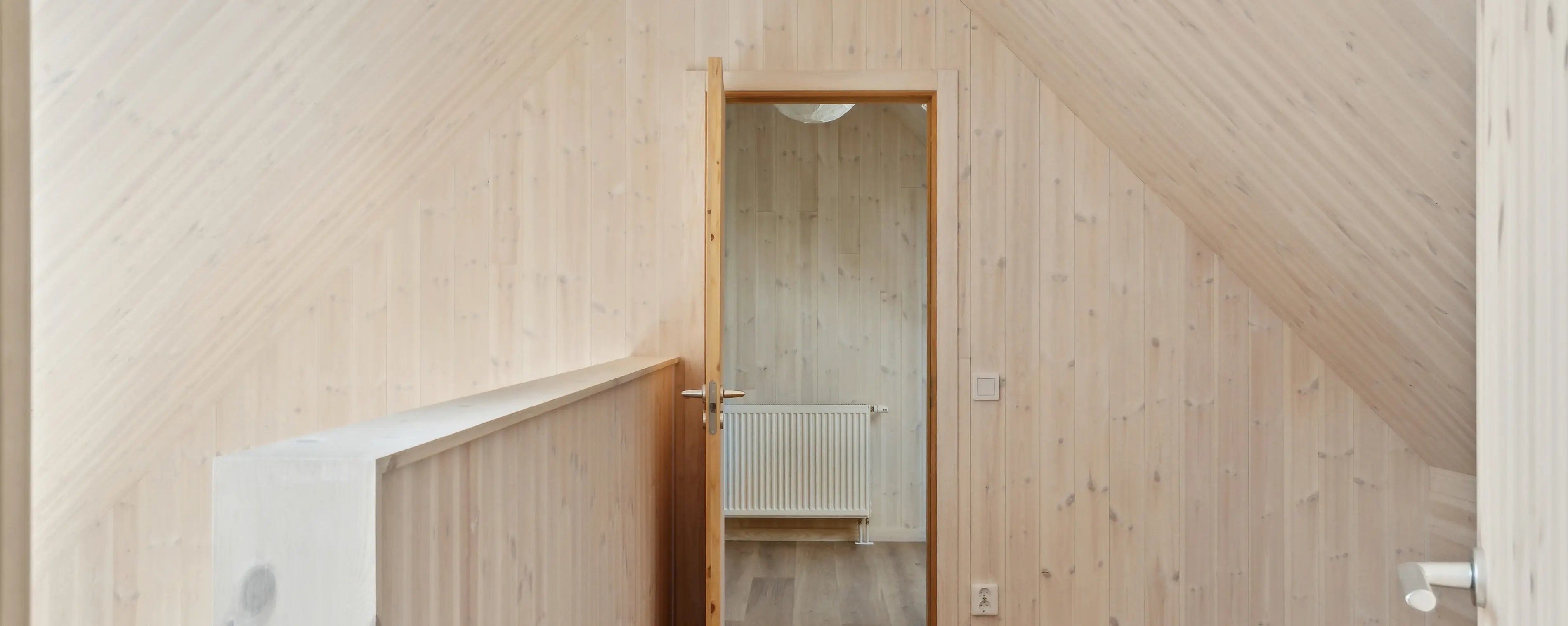 Narrow hallway with light wooden walls and sloped ceiling, leading to a small room with a white radiator.