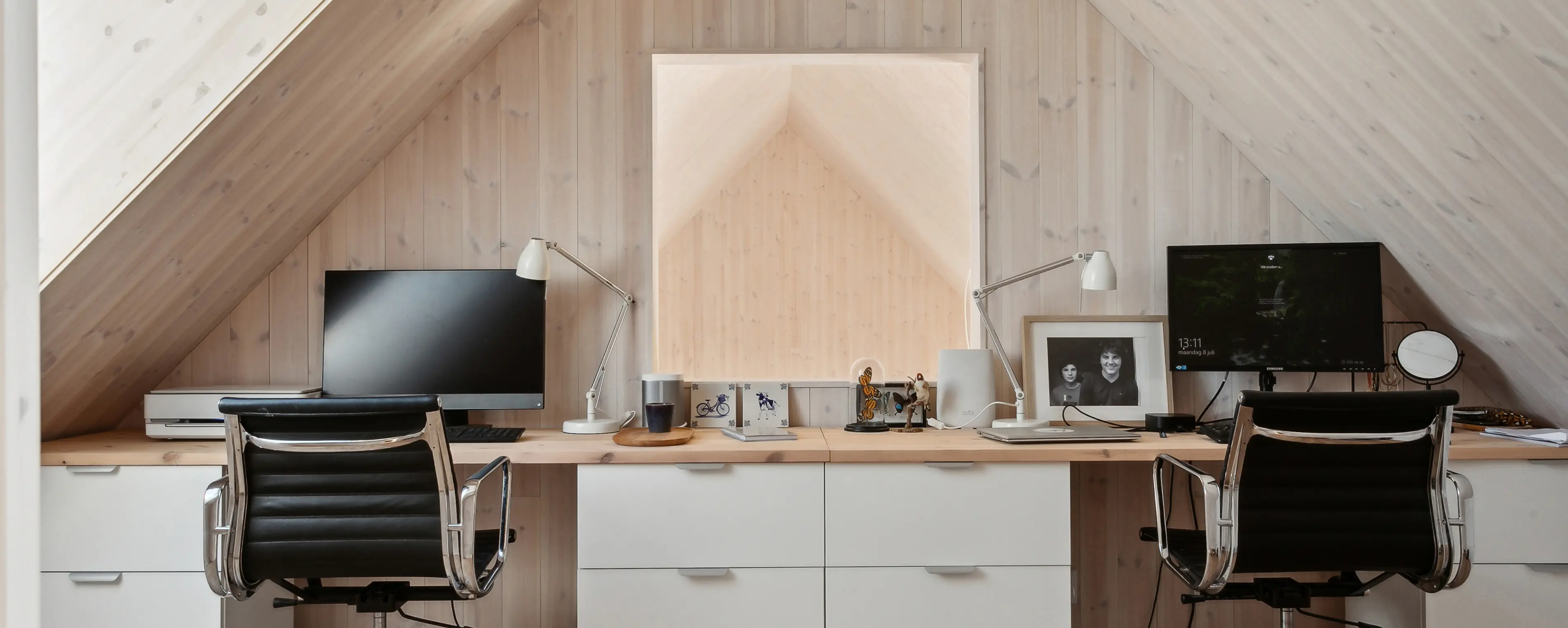 Modern attic home office with two desks, dual monitors, white drawers, and slanted wooden ceilings.