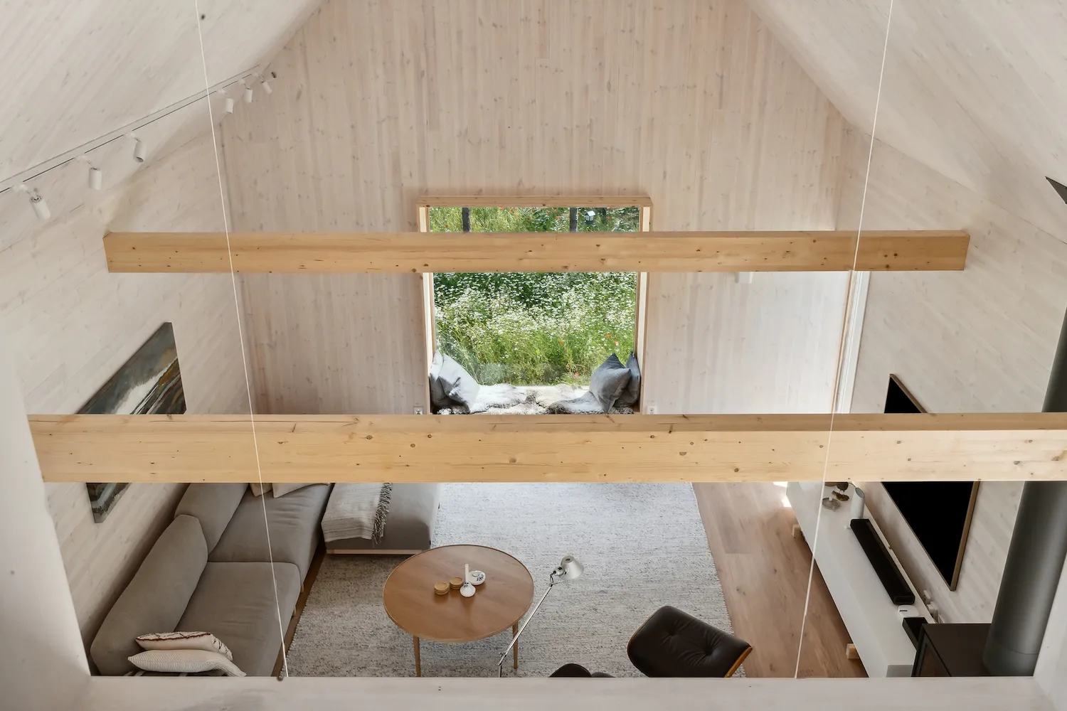 Interior of the Arvesund house model Dalasen, with high wooden ceilings, exposed beams, large window opening to greenery, and a cozy living area with sofa and round table.