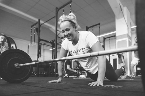 Sportlerin lacht während eines CrossFit-Trainings, liegt nach einer Übung neben einer Langhantel auf dem Boden.