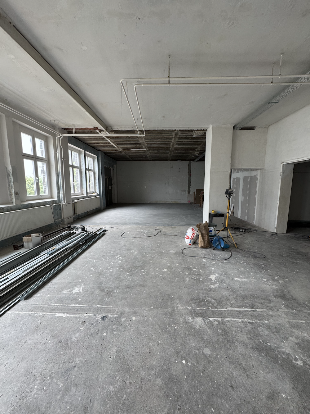 Leerer Raum im Rohbauzustand mit großen Fenstern und Baumaterialien am Boden.