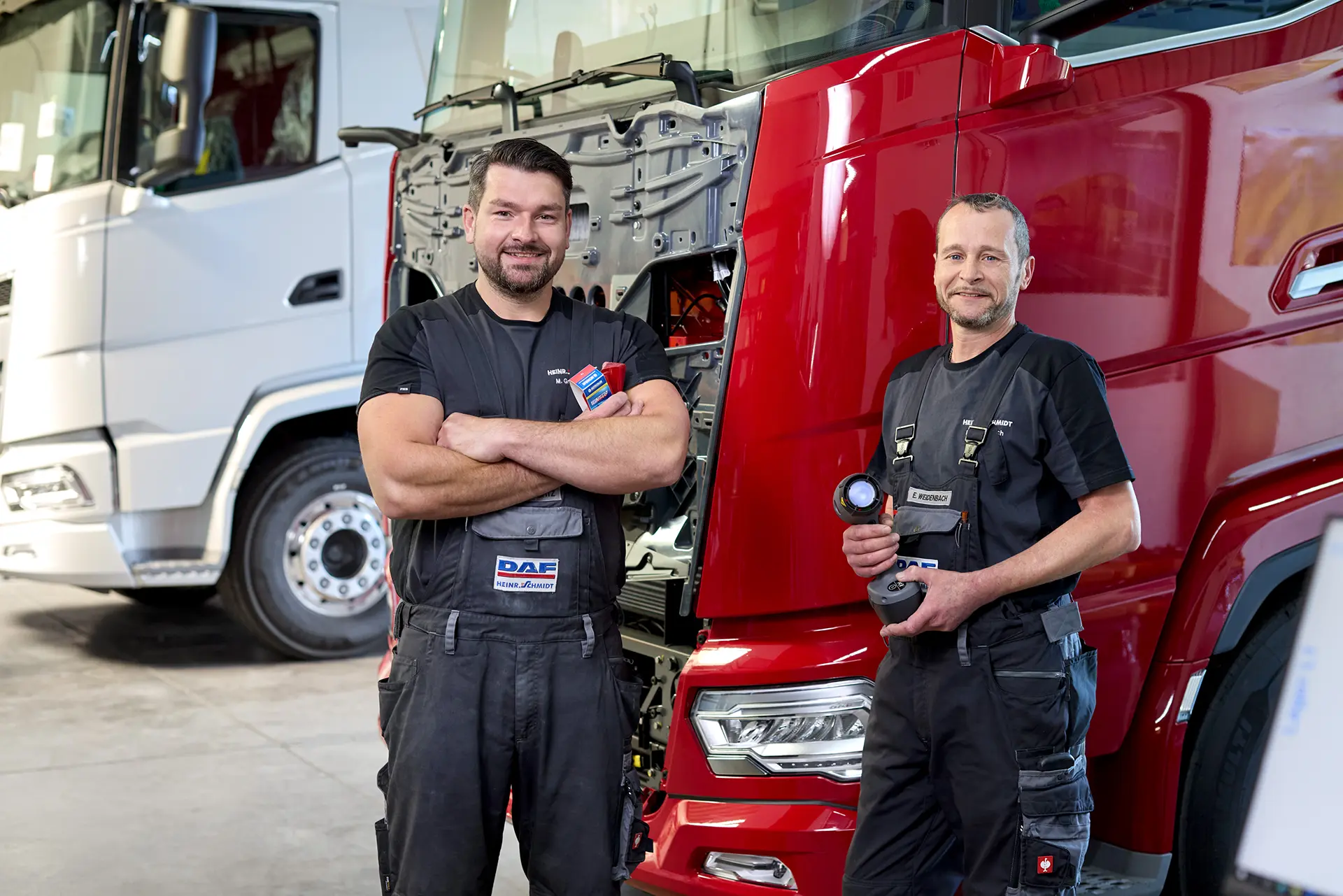 Zwei DAF-Mechaniker in der Werkstatt von Heinr. Schmidt in Paderborn stehen vor einem roten Lkw und lächeln in die Kamera.